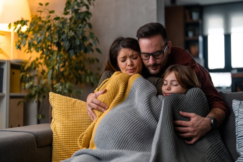 Why Isn’t My Furnace Heating My Entire Home? Photo of a family of three freezing in their home, huddled in a blanket to keep warm.